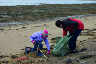 An adult and a child clean the strandline on a sandy beach with a litter picker