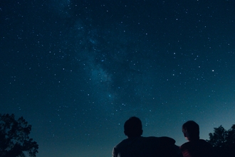 Image showing a couple in silhouette under the stars. They are just off-centre and there is a tree to their left and one to their right.