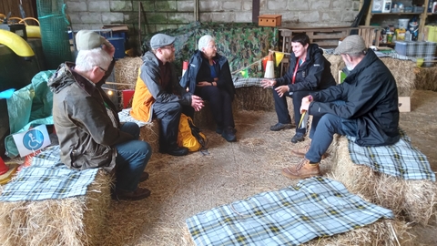 Six people are in a barn sat on hay bales in a circle having a discussion. 