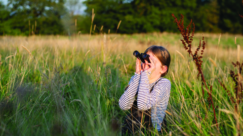 Girl stands amongst the grass looking through binoculars
