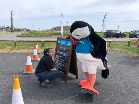 A photograph of Joy writing on a chalkboard with Cliff the Puffin