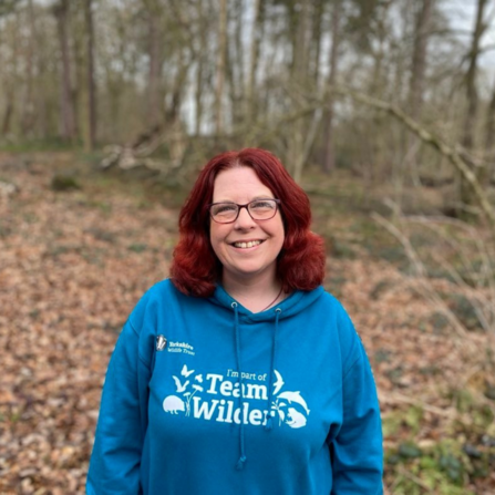 Woman with red hair in teal #TeamWilder hoodie stood in woodland smiling facing the camera