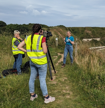 Two women with a camera in high vis stand to the left, whilst a staff member in a blue t shirt stands to the right. The group are on a clifftop. 