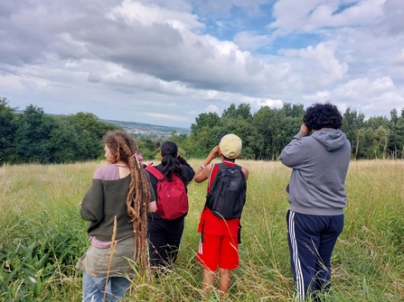 Group of youths out in nature using binoculars. They are standing in long grass in an open green space with trees and hills in the distance.