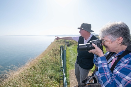 Two people looking out to see, one through a short telescope and the other is pointing. It's a sunny day and they are stood on the edge of a coastline.