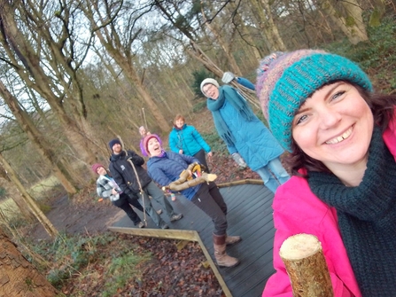 Selfie of a group of people walking along a boardwalk in a nature reserve carrying wood and small tree branches that they have just chopped.