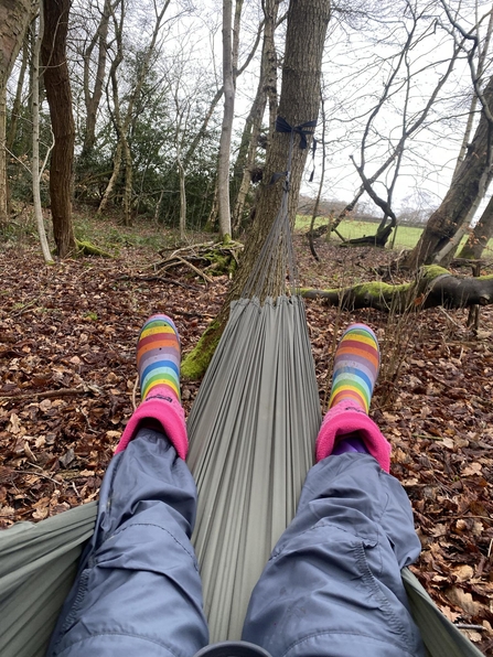 View from a person sat in a hammock in the woods, you can see their legs and rainbow wellies and some of the forest