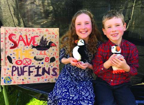 Sophie and Theo sat next to a 'Save the puffins' sign, both holding a cuddly puffin