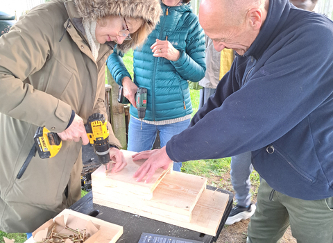 Woman making bird and bat boxes on a table outside. She is being assisted by a man and another woman is watching on and smiling.