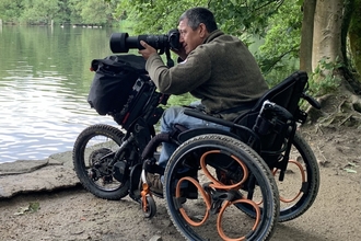 Colour photo of Peter Lau in his wheelchair taking photos with a long lens at a nature reserve 