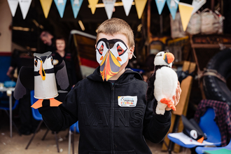 A child wearing a puffin mask holding a puffin figure he's made.