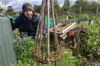 Man crouched down in an allotment. In the forefront of the shot is a wigwam for plants and veg to grow up.