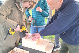 Woman making bird and bat boxes on a table outside. She is being assisted by a man and another woman is watching on and smiling.