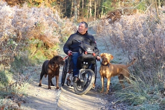 Man in a 3-wheeler all terrain mobility scooter on a nature reserve with 2 dogs on short leads.