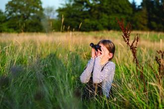 Girl stands amongst the grass looking through binoculars