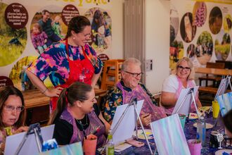 A group sat around a long table with easels in a painting class. There is a teacher standing behind admiring a student's work and everyone is looking toward's the man's work and smiling.
