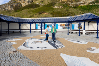 Two people stand in the centre of the paved area on top of the mosaic, with the covered area behind.