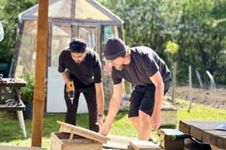 Two men build a wooden structure outdoors on a sunny day. One man uses a power drill while the other inspects the structure. A garden and a greenhouse are visible in the background.