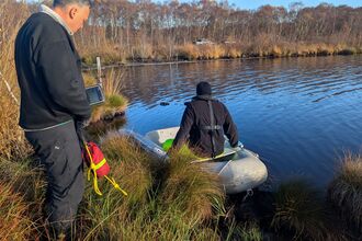 person on a small dingy on the water at the edge. Another man is stood on the bank monitoring.