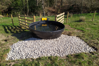 A large black circular trough of water sites above a gravelled square area cut out of the grass