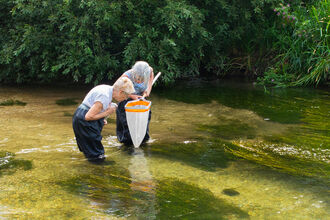 Two women wading in a shallow, clear stream using a net to collect water samples, with lush greenery surrounding the riverbank.