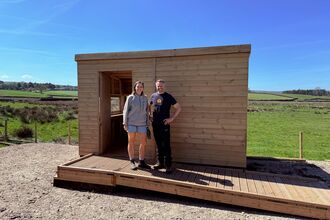 Two people stand in front of a newly built wooden bird hide on a sunny day. The hide is rectangular, with an open doorway and built-in wooden deck. A woman in a grey zip-up jacket and denim shorts stands left of the door, and a man in a black t-shirt and dark pants stands right. They are positioned against a backdrop of green fields and a blue sky.