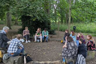 Group of people sat in a circle on logs in a woodland listening to someone read poetry from notebook. 