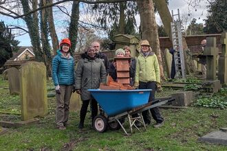 A group of six people stand in a cemetery next to a blue wheelbarrow filled with wooden planks and a birdhouse.