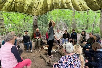 Group of people sat in circle on logs around unlit firepit watching person in centre instruct them on breathwork