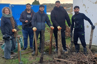 A group of five people stand in a muddy yard outdoors next to a pile of branches and wooden stakes, with a blue utility structure and a weathered building in the background.