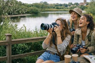 group of friends birdwatching and laughing, one holds binoculars to their eyes and there are some coffee cups and bird id book on the table. Behind them is a nature reserve setting with a wooden fence and a body of water behind it.