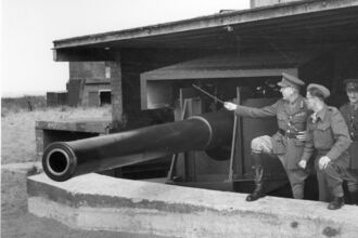 Men in military uniforms stand next to a large coastal artillery gun within a protective concrete emplacement and wooden structure, with one man pointing at the gun.