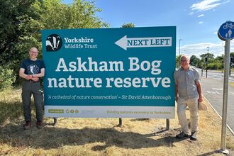A large blue sign with 'Askham Bog nature reserve' written on it has been unveiled. Two people stand either side of it - looking very happy!