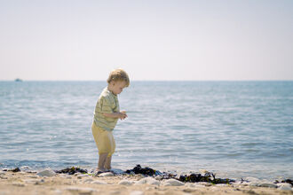 A blond-haired toddler wearing a striped shirt and yellow shorts stands at the edge of the ocean, looking down at something in their hands. The water is calm and blue, with a gentle surf lapping at the sandy shore. In the distance, a small boat is visible on the horizon under a clear, pale sky.
