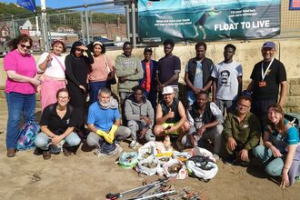 Group of volunteers posing on Scarborough beach after a litter pick, with bags of collected rubbish and litter-picking tools arranged in front. The group stands and kneels in front of a sea wall with the harbour and hillside visible in the background on a sunny day.