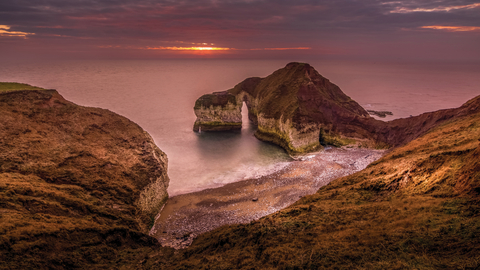 A cliff that juts out into the sea that looks like a dinosaur drinking. The sun is setting over the sea