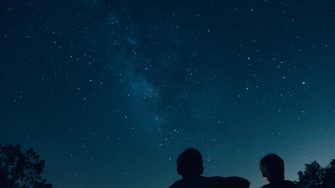 Image showing a couple in silhouette under the stars. They are just off-centre and there is a tree to their left and one to their right.