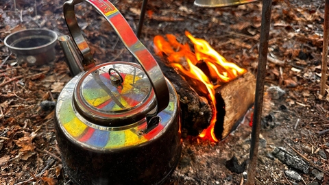 Campfire and teapot which has a rainbow reflection on it. They are laid on the woodland floor for forest school