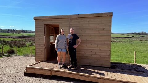 Two people stand in front of a newly built wooden bird hide on a sunny day. The hide is rectangular, with an open doorway and built-in wooden deck. A woman in a grey zip-up jacket and denim shorts stands left of the door, and a man in a black t-shirt and dark pants stands right. They are positioned against a backdrop of green fields and a blue sky.