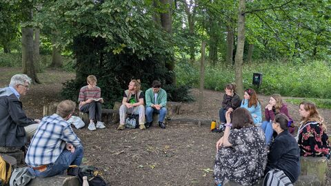 Group of people sat in a circle on logs in a woodland listening to someone read poetry from notebook. 