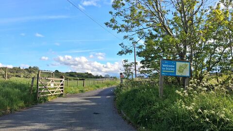 A scenic pathway leads into a green field with a wooden gate on the left and a "Welcome to Stirley" sign with a map on the right, under a bright blue sky with fluffy clouds.