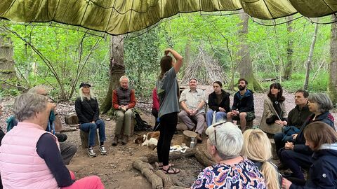 Group sit on logs in circle facing coach guiding them through breathing exercise under tarp