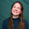 A young woman with long, brown hair smiles broadly at the camera, wearing a dark blue turtleneck sweater against a teal background with diagonal shadow patterns.