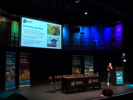 Rosie stands at a podium at our AGM on the right with posters and a powerpoint behind her