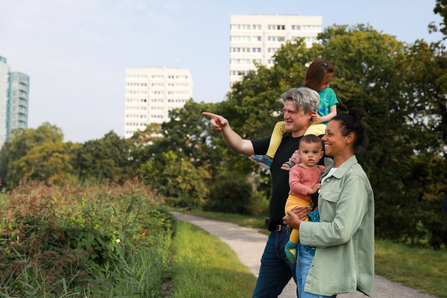 A family of four are at a nature reserve with buildings visible in the background. The father is pointing to show his partner and children something in the distance