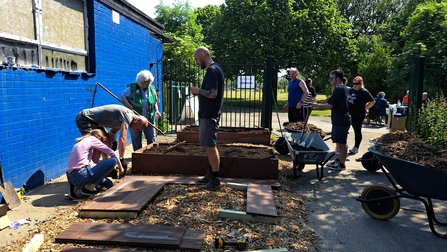 A group of people in the centre of the image are knelt around several wooden boards building a raised border to the right of a blue building