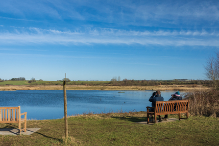 Two people sitting on a bench with binoculars looking out across the wetland lake on a nature reserve on a sunny cold day