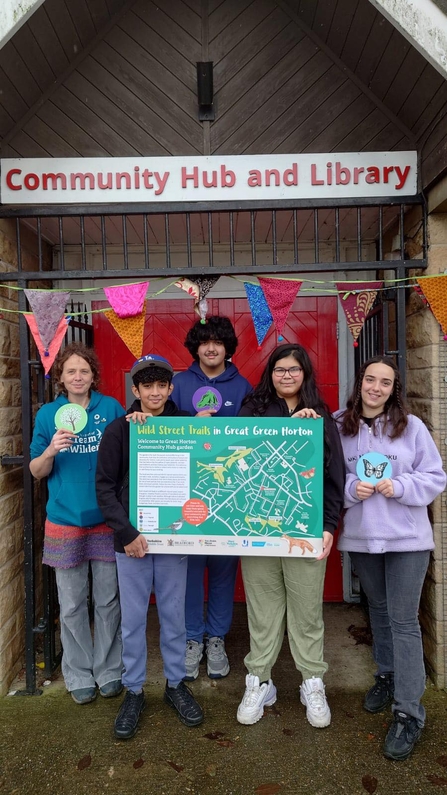 Group of youths holding a board depicting a map of their wild street trails that they have designed. They are stood under a sign saying Community Hub and Library