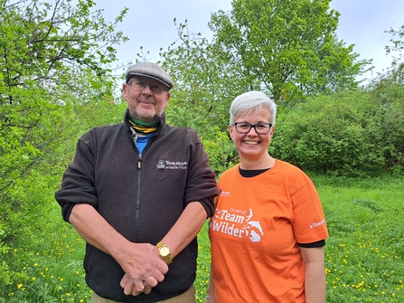 Man and woman stood facing the camera smiling, standing on the grounds of a rugby club that is supporting nature