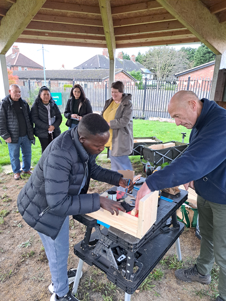 Man making some bird boxes assisted by another man on a table outside. In the rear of the shot are 4 people watching. 